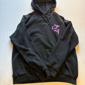 Svart hoodie med grafiskt tryck - Svart hoodie med stor huva och dragsko. Framtill finns ett lila och vitt grafiskt tryck på bröstet och på ryggen ett stort, färgstarkt tryck i lila, vitt och blått med textdetaljer. Mjuk insida och klassisk känguruficka framtill. Perfekt för dig som gillar streetwear.