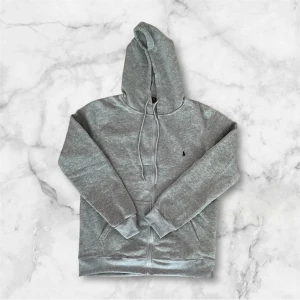 Grå hoodie från Polo Ralph Lauren - Snygg grå hoodie från Polo Ralph Lauren med dragkedja och huva. Tröjan har broderad logga på bröstet, dragsko i huvan och två fickor framtill. Perfekt för en chill och stilren look.