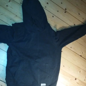 Svart hoodie från 157 med ficka - Svart hoodie från 157 med klassisk känguruficka framtill och stor huva. Tröjan är i mjuk bomull och har långa ärmar. Perfekt för chill dagar eller när du vill ha något bekvämt och enkelt.