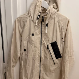 Beige jacka med huva från Mouli - Snygg beige jacka från Mouli med huva, svarta detaljer och dragkedja i sned design. Jackan har flera tryckknappar, svart bröstficka och dragsko i huvan. Perfekt för dig som gillar stilren och funktionell look. Storlek M.