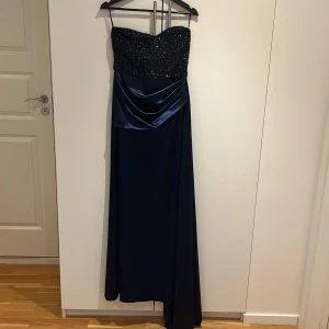 Marinblå glittrig långklänning - Elegant marinblå långklänning med glittrig överdel och smala axelband. Klänningen har en snygg drapering i satin över midjan och en figurnära passform upptill. Perfekt för dig som vill sticka ut med en lyxig och stilren look.