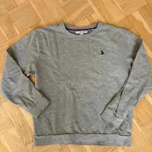 Grå sweatshirt från U.S. Polo Assn. - En stilren grå sweatshirt från U.S. Polo Assn. med rund halsringning och broderad logga på bröstet. Tröjan har långa ärmar och ribbade muddar vid ärmslut och nederkant. Perfekt för en avslappnad och snygg look.
