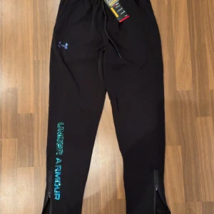 Svarta träningsbyxor Under Armour M - Svarta träningsbyxor från Under Armour i loose fit med elastisk midja och snörning. Byxorna har dragkedjor vid bensluten och logga i blått och grönt längs benet. Perfekta för träning eller chill, tillverkade i lätt syntetmaterial.