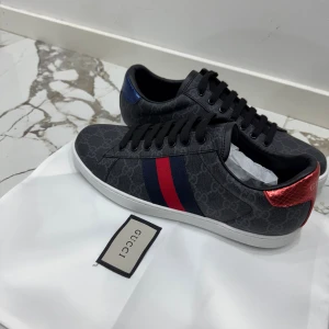 Gucci sneakers med GG-mönster - Snygga Gucci sneakers i mörkgrått med GG-monogram över hela skon. Klassiska röda och blå ränder på sidan, samt metallicröd detalj på ena hälen och metallicblå på den andra. Skorna har svarta snören och vit sula. Perfekt för dig som vill sticka ut med lyxig streetstyle.