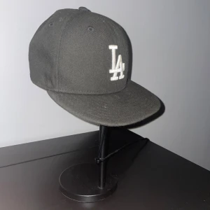 Svart LA Keps - Säljer en svart New Era 59FIFTY keps. Klassisk flat peak och MLB-logga baktill. Kepsen är tillverkad i 100% polyester och har en stilren, sportig look som passar perfekt till streetwear. Storlek 7