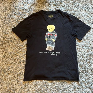Svart Polo Bear t-shirt från Ralph Lauren - Svart t-shirt från Polo Ralph Lauren med ikoniskt Polo Bear-tryck på bröstet. Björnen har en stickad tröja med mönster och jeans, vilket ger en cool och lekfull vibe. T-shirten är i mjuk bomull och har klassisk passform med korta ärmar.