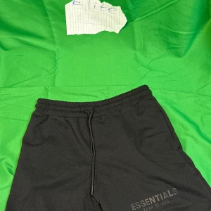 Svarta Essentials Fear of God mjukisbyxor - Svarta mjukisbyxor från Essentials Fear of God med elastisk midja och snörning. Byxorna har en stilren look med logga tryckt på benet och är tillverkade i mjukt material för maximal komfort. Perfekta för en avslappnad och trendig stil.Liknande modell som originalet!