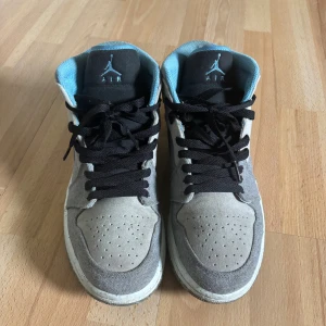 Nike Air Jordan 1 grå/blå sneakers - Nike Air Jordan 1 sneakers i grått och ljusblått med svart snörning och hög siluett. Ovandel i textil och mocka, blå meshfoder och klassisk Jordan-logga på plösen. Säljes med vita toe guards för extra skydd. Perfekta för dig som gillar streetwear och ikoniska sneakers. Pris kan diskuteras. Skriv om ni har någon fråga.