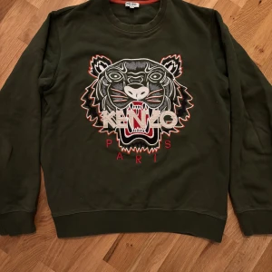 Kenzo sweatshirt - Säljer en grön sweatshirt från Kenzo med ikonisk tigerbrodyr och Kenzo Paris-text framtill. Tröjan har rund halsringning, långa ärmar och ribbade muddar. Perfekt statement-plagg för dig som vill sticka ut.
