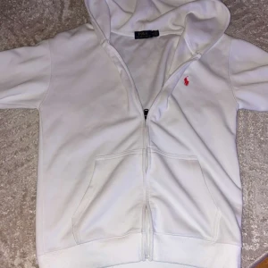 Vit hoodie från Polo Ralph Lauren - Vit hoodie från Polo Ralph Lauren med dragkedja och huva. Klassisk design med röd broderad logga på bröstet och stora fickor framtill. Tillverkad i mjuk bomull, perfekt för en clean och stilren look. säljer den då för när jag köpte den var den för liten 