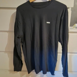 Svart långärmad tröja från Vans - En stilren svart långärmad tröja från Vans med liten vit logga på bröstet. Tröjan har rund halsringning och är tillverkad i mjuk bomull, perfekt för en avslappnad streetwear-look. Storlek M