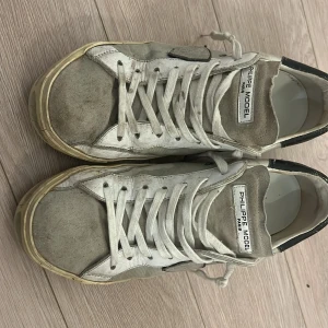Philippe Model vita sneakers med svarta detaljer - Säljer ett par Philippe Model sneakers i vitt och grått med svarta detaljer på hälen. Skorna har snörning, rund tå och platt sula. Ovandelen är i en mix av skinn och mocka för en cool, urban look. Lite småsönder där bak anledning till låga pris