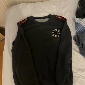 Grå Dolce & Gabbana sweatshirt - Snygg grå sweatshirt från Dolce & Gabbana med broderad detalj på bröstet och röda axelpatchar med guldbroderi. Tröjan har rund halsringning och långa ärmar, perfekt för dig som vill sticka ut med exklusiva detaljer.
