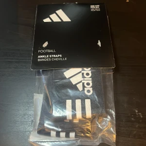 Adidas svarta ankelband fotboll - Svarta ankelband från Adidas med klassiska vita ränder och logga. Perfekta för fotbollsträning och matcher. Elastiskt material som sitter skönt runt vristen och håller benskydden på plats.