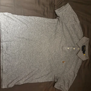 Grå pikétröja Polo Ralph Lauren - Snygg grå pikétröja från Polo Ralph Lauren i slim fit-modell. Klassisk krage, tre knappar framtill och den ikoniska broderade loggan på bröstet. Perfekt för en clean och stilren look.