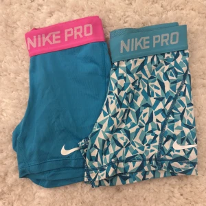 Nike Pro blå träningstights shorts - Två par Nike Pro shorts i blå toner, ett par helblå med rosa resår och ett par med blått geometriskt mönster och blå resår. Båda har Nike-logga och är perfekta för träning. Sitter tight och har elastisk midja för bästa komfort.