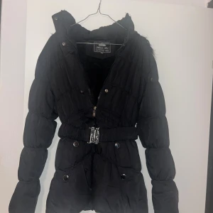 Svart pufferjacka med bälte Hua Li  - Svart pufferjacka från Hua Li i storlek XXL. Jackan har en fluffig huva med fuskpäls, markerad midja med brett bälte och silvriga detaljer. Dragkedja framtill och tryckknappar, samt mjukt foder på insidan. Perfekt för kalla dagar och riktigt snygg siluett.