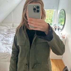 Olivgrön parkajacka med snörning - Snygg olivgrön jacka med dragsko i midjan och två stora bröstfickor. Jackan har dragkedja framtill och raka armar. Den är i mycket bra skick