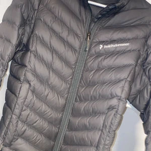 Grå dunjacka Peak Performance S - Snygg grå dunjacka från Peak Performance i storlek S. Jackan har diskret logga på bröstet, dragkedja framtill och klassisk quiltad design. Perfekt för kyliga dagar och enkel att matcha med allt. Lätt och smidig att bära med sig. Som ny 