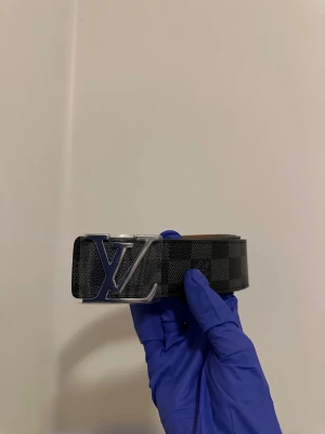 L och V rutig svart bälte - Snyggt L och V bälte med klassiskt L och V-spänne i silver. Bältet har ett rutigt mönster i svart och grått, tillverkat i skinn med en lyxig känsla. Perfekt accessoar för att lyfta din outfit med exklusiv vibe och stil.