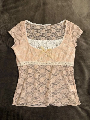 Rosa spetsblus med rosett - Supersöt rosa  blus i spets med blommigt mönster och korta ärmar. Toppen har en dekorativ rosett framtill och en v-ringad hals med vit rynkad detalj. Längd :45 cm,  midja:60 cm