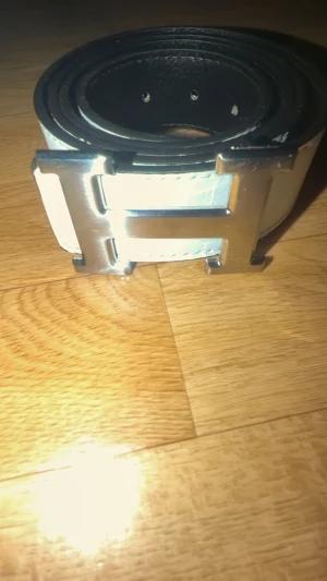 Vit skärp med silver H-spänne - Snyggt vitt skärp med ett stort H-format spänne i silverfärgad metall. Skärpet är tillverkat i skinn med en slät och lätt texturerad yta. Passar perfekt för att ge din outfit en clean och stilren touch. Bredden är rejäl och spännet är riktigt iögonfallande.