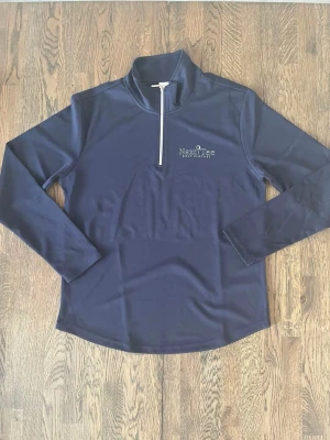 Marinblå golftröja från Next Tee - Snygg marinblå långärmad tröja från Next Tee Golf Clothes med half zip framtill. Tröjan har en stilren design med vit dragkedja och diskret logga på bröstet. Perfekt för golf eller chill dagar. Materialet är polyester och passformen är normal.