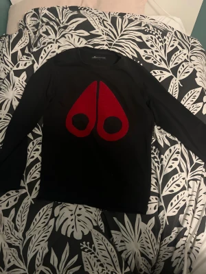 Svart tröja Moose Knuckles med rött tryck - Svart långärmad tröja från Moose Knuckles i storlek S. Framsidan har ett stort rött Moose Knuckles-tryck och på ärmen finns en svart patch med loggan. Tröjan har rund hals och är gjord i mjukt bomullsmaterial, perfekt för chill dagar.