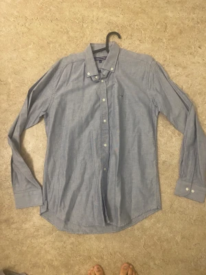 Blå skjorta från Tommy Hilfiger - Klassisk blå skjorta från Tommy Hilfiger med button down-krage och små logodetaljer på bröstet. Skjortan har långa ärmar, vita knappar och är tillverkad i mjuk bomull. Perfekt för en clean och stilren look.