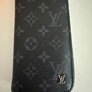 Svart plånbok Monogram - Snygg svart plånbok med klassiskt monogrammönster och silverfärgad logga. Plånboken har dragkedja runtom, flera kortfack och fack för sedlar. Perfekt för dig som gillar lyxiga accessoarer med stilren design.