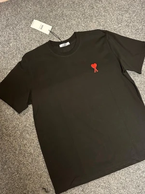 Svart t-shirt från Ami Paris  - Svart t-shirt från Ami Paris i mjuk bomull med klassisk rund hals och korta ärmar. På bröstet sitter Ami:s ikoniska röda hjärta broderat. Enkel och stilren design som lyfter din outfit med en snygg detalj.