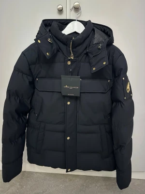 Svart dunjacka Moose Knuckles L - Säljer en svart dunjacka från Moose Knuckles i storlek L. Jackan har guldiga detaljer, dragkedja och knappar, samt flera stora fickor och en avtagbar huva. Insidan har mönstrat foder och logotypband. Perfekt för kalla dagar och riktigt snygg streetstil. Prislappen sitter på 