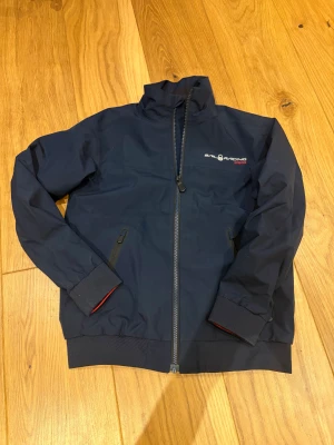 Marinblå vindjacka från Sail Racing - Snygg marinblå vindjacka från Sail Racing med dragkedja framtill och två fickor med dragkedja. Jackan har hög krage och diskret logga på bröstet. Tillverkad i slitstark polyester och perfekt för blåsiga dagar.