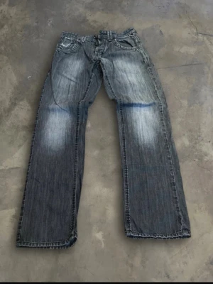 Jeans Angel Litrico grå tvätt - Säljer ett par Angel Litrico jeans i grå tvätt. Jeansen har klassiska fem fickor, slitna detaljer vid fickorna och en lågmidjad design. Materialet är robust denim och byxorna har knapp framtill.