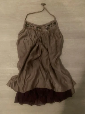  halterneck topp med broderi - Säljer en halterneck topp med broderade detaljer upptill och volangkant i mörklila nedtill. Toppen har en lös passform och är tillverkad i ett tunt, glansigt material som ger en snygg drapering. Perfekt för dig som gillar unika detaljer och boho-vibe.