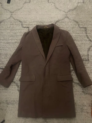 Brun rock från J.Lindeberg - Snygg brun rock från J.Lindeberg med klassisk krage och två fickor framtill. Insidan är quiltad för extra komfort och värme. Perfekt för dig som vill ha en stilren och tidlös look. Materialet känns mjukt och har en exklusiv känsla.