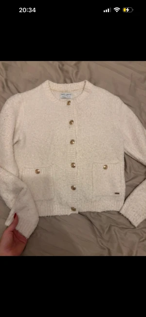 Vit kofta från Holly & White - Superfin vit kofta från Holly & White med guldfärgade knappar och två fickor framtill. Koftan är stickad och har en mjuk, fluffig känsla. Rund halsringning och långärmad modell som är perfekt för kyliga dagar.