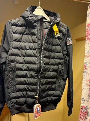 Svart lättviktsjacka Parajumpers S - Svart superlätt pufferjacka från Parajumpers med huva, quiltad framsida och ärmar i slätare material. Jackan har dragkedja, två fickor med dragkedja och ikonisk gul detalj vid axeln. Märkespatch på ärmen och snygg passform.