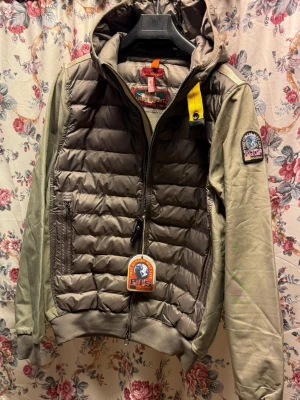 Parajumpers grön pufferjacka med huva - Snygg pufferjacka från Parajumpers i olivgrönt och grått med quiltad framsida, huva och gul detalj vid halsen. Jackan har dragkedja, ribbade muddar och patch på ärmen. Perfekt för dig som vill ha en cool och funktionell jacka med streetkänsla.