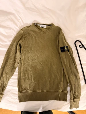 Olivgrön tröja från Stone Island - Säljer en olivgrön långärmad tröja från Stone Island med den klassiska loggan på vänster ärm. Tröjan har rund halsringning och ribbade muddar vid ärmslut och nederkant. Perfekt för dig som gillar streetwear och snygga detaljer.