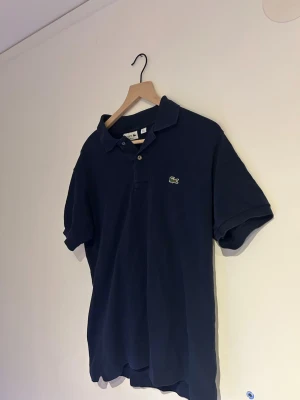 Marinblå pikétröja från Lacoste - Säljer en klassisk marinblå pikétröja från Lacoste med den ikoniska gröna krokodilloggan på bröstet. Hör av er vid frågor eller intresse