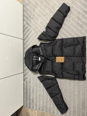 Svart pufferjacka från Burberry - Säljer en svart pufferjacka från Burberry med avtagbara ärmar och huva. Jackan har gråmelerat foder med ränder, dragkedjor på fickorna och en tydlig Burberry-patch på ärmen. Perfekt för kalla dagar och riktigt snygg streetstil.