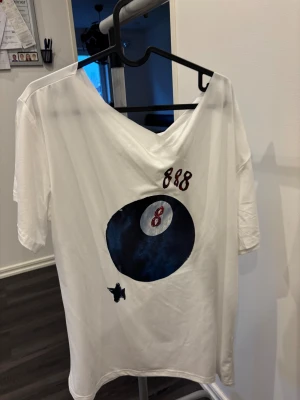 Vit t-shirt med 8-ball tryck - Vit t-shirt med v-ringning och tryck av en svart biljardboll (8-ball) framtill. Siffran 848 är tryckt ovanför motivet. T-shirten har korta ärmar och en avslappnad passform, perfekt för en cool och avslappnad stil.