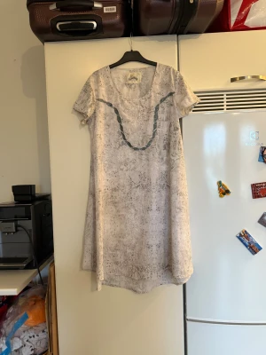 Ljus kortklänning från Fira med glitter - Supersöt ljus kortklänning från Fira med diskret grått mönster och glittriga detaljer. Klänningen har korta ärmar, rund halsringning och en dekorativ flätad detalj framtill. Perfekt för dig som gillar unika och bekväma plagg. Storlek M. Mycket fint skick, jag hittar inga synliga anmärkningar eller defekter. 