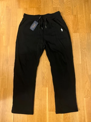 Svarta mjukisbyxor Polo Ralph Lauren M - Svarta mjukisbyxor från Polo Ralph Lauren med dragsko i midjan och klassisk broderad logga på höften. Byxorna har normal passform och är tillverkade i mjukt bomullsmaterial. Perfekta för en chill och avslappnad stil.