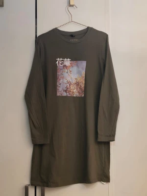 Khakigrön långärmad klänning DeFacto - Khakigrön långärmad klänning från DeFacto i bomull med ett tryck framtill som visar ett foto med japanska tecken. Rak passform och rund halsringning, perfekt för en chill och avslappnad look.