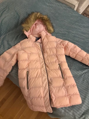 Ljusrosa pufferjacka från Vinson Polo Club - Säljer en ljusrosa pufferjacka från Vinson Polo Club med huva och fejkpäls. Jackan har två fickor med dragkedja, quiltad design och guldiga detaljer. Perfekt för kalla dagar och har en snygg, modern look som sticker ut. Väldigt bra skick,använd ett par gånger därför högt pris, köpte den för ungefär 800kr.