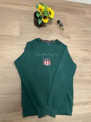 Grön sweatshirt från GANT - Snygg grön sweatshirt från GANT med broderad logga och emblem på bröstet. Tröjan har rund hals och långa ärmar, samt ribbade muddar vid ärmslut och nederkant. Perfekt för dig som gillar klassisk stil med en sportig touch.