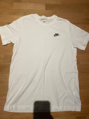 Vit Nike t-shirt med logga - Säljer en clean vit t-shirt från Nike med klassisk svart logga på bröstet. T-shirten har rund hals och korta ärmar, perfekt för en chill och sportig stil. Materialet är mjuk bomull som känns skönt mot huden.