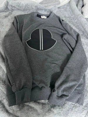 Grå sweatshirt från Moncler - Mörkgrå sweatshirt från Moncler med stort svart och vitt tryck på bröstet. Tröjan har rund hals, ribbade muddar och är tillverkad i mjuk bomull. Perfekt för en chill och stilren look.